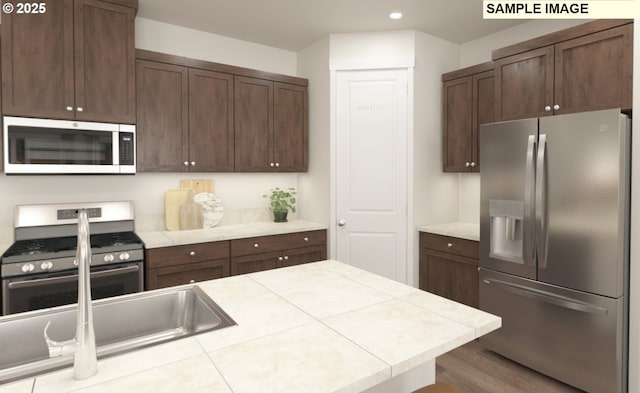 kitchen featuring tile countertops, wood finished floors, recessed lighting, dark brown cabinetry, and appliances with stainless steel finishes