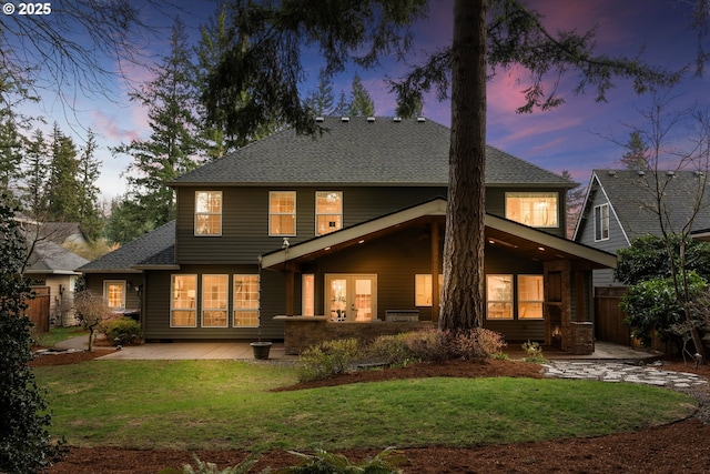 back of house at dusk with a lawn, a patio, fence, french doors, and roof with shingles