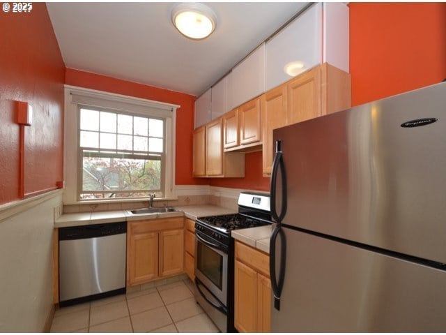 kitchen featuring tile countertops, light tile patterned flooring, stainless steel appliances, a sink, and light brown cabinetry