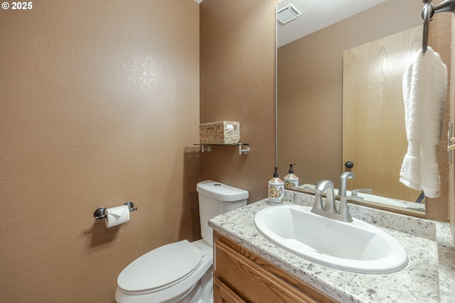 half bathroom featuring vanity, a textured wall, visible vents, and toilet