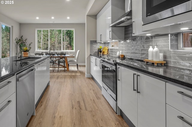 kitchen featuring wall chimney range hood, appliances with stainless steel finishes, a sink, light wood-style floors, and white cabinetry