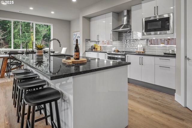 kitchen featuring wall chimney range hood, a sink, built in microwave, white cabinetry, and a kitchen bar