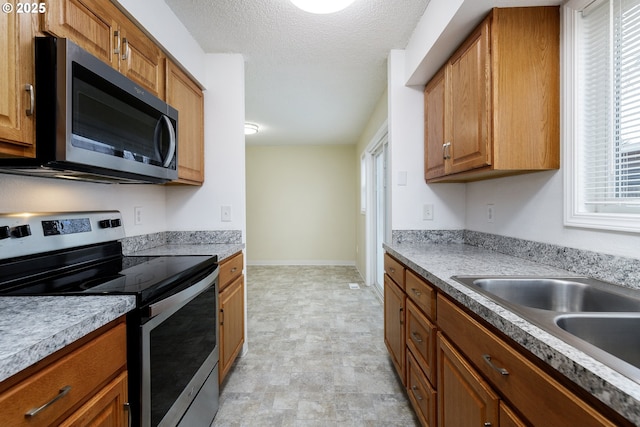 kitchen featuring brown cabinetry, baseboards, a sink, appliances with stainless steel finishes, and a textured ceiling