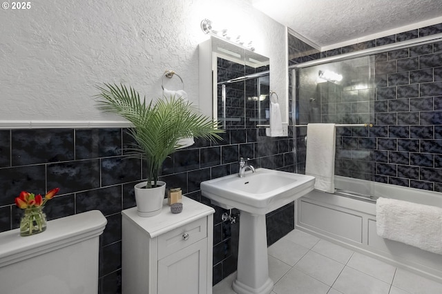 bathroom with combined bath / shower with glass door, tile patterned flooring, toilet, a textured ceiling, and tile walls