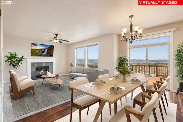 dining space with a textured ceiling, a tiled fireplace, ceiling fan with notable chandelier, baseboards, and wood finished floors