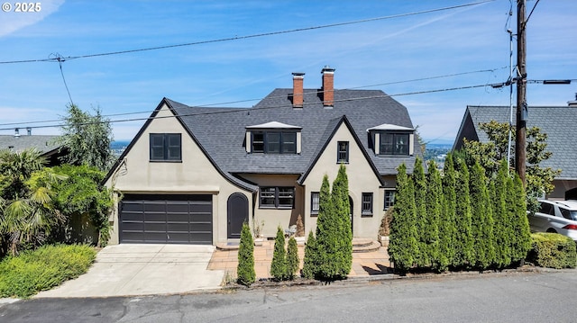 tudor-style house with a garage, roof with shingles, driveway, and stucco siding