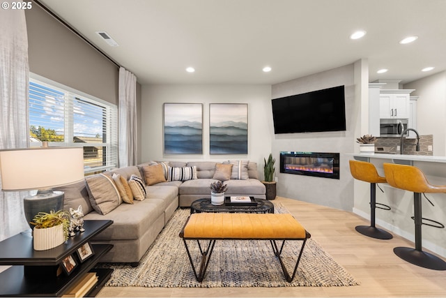 living area featuring light wood-style floors, a glass covered fireplace, visible vents, and recessed lighting