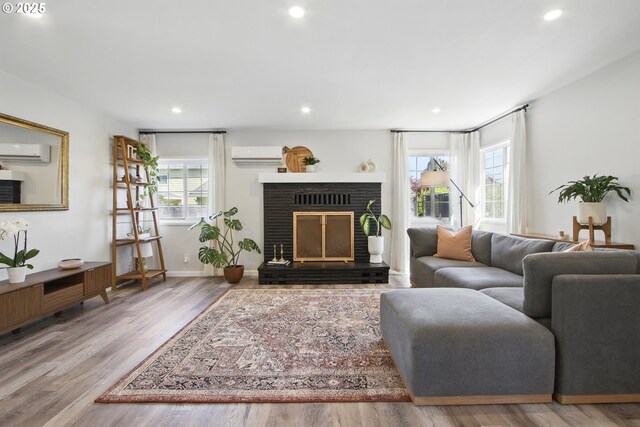 living area with a wall mounted air conditioner, wood finished floors, a brick fireplace, recessed lighting, and baseboards