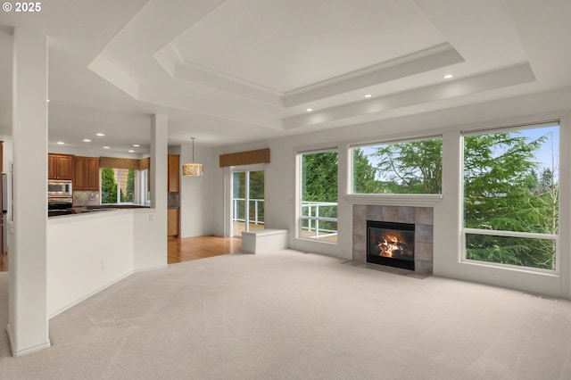 unfurnished living room with a tiled fireplace, a tray ceiling, light carpet, and a healthy amount of sunlight
