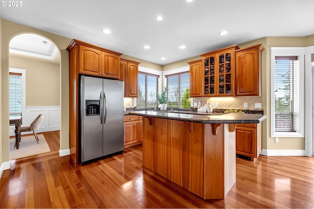 kitchen with arched walkways, wood finished floors, a kitchen breakfast bar, stainless steel refrigerator with ice dispenser, and glass insert cabinets