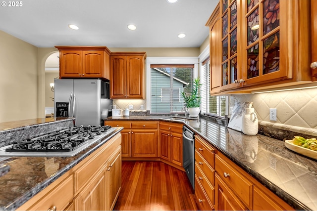 kitchen with arched walkways, glass insert cabinets, appliances with stainless steel finishes, dark stone countertops, and a sink