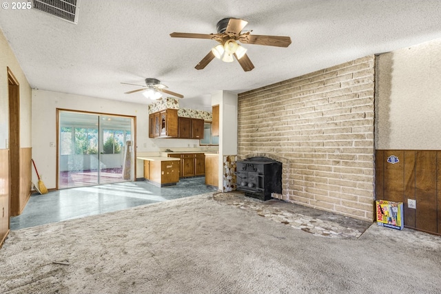 unfurnished living room with wood walls, a textured ceiling, a wood stove, carpet flooring, and ceiling fan