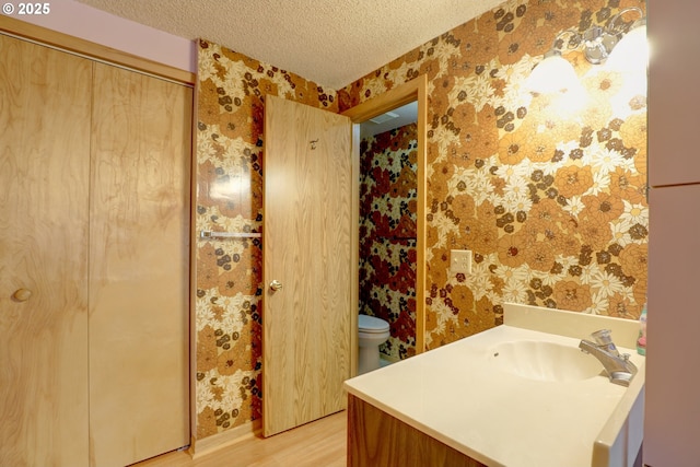 bathroom featuring wallpapered walls, toilet, wood finished floors, a textured ceiling, and vanity