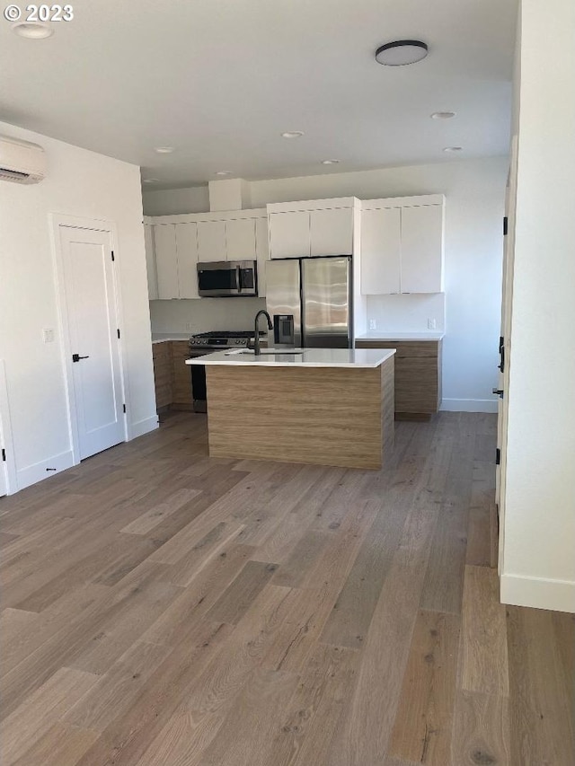 kitchen with an AC wall unit, an island with sink, white cabinets, and appliances with stainless steel finishes