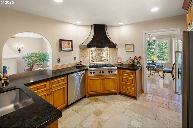 kitchen featuring brown cabinetry, premium range hood, stainless steel appliances, tasteful backsplash, and stone tile flooring
