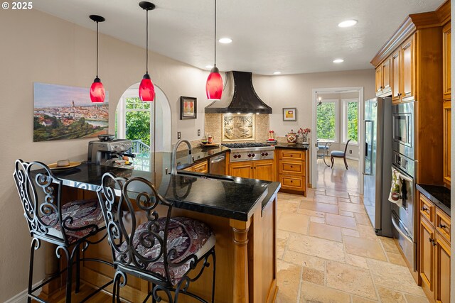 kitchen featuring wall chimney range hood, brown cabinetry, appliances with stainless steel finishes, dark countertops, and stone tile floors