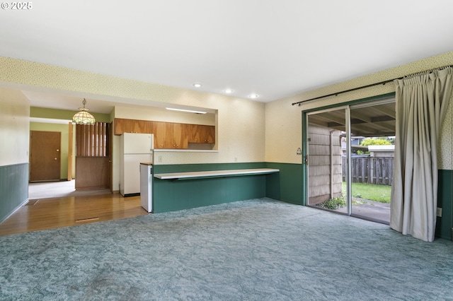 kitchen with light colored carpet, freestanding refrigerator, a peninsula, brown cabinetry, and a chandelier