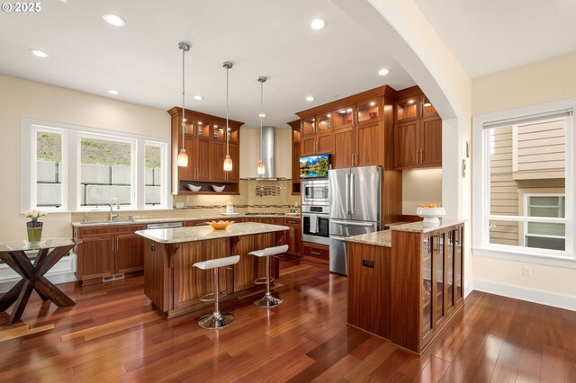 4018 NW Riggs Dr, Portland, OR 97229 - kitchen featuring light stone counters, wall chimney range hood, brown cabinetry, stainless steel appliances, and arched walkways