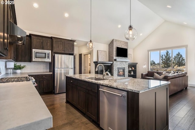 kitchen with appliances with stainless steel finishes, a stone fireplace, pendant lighting, an island with sink, and sink