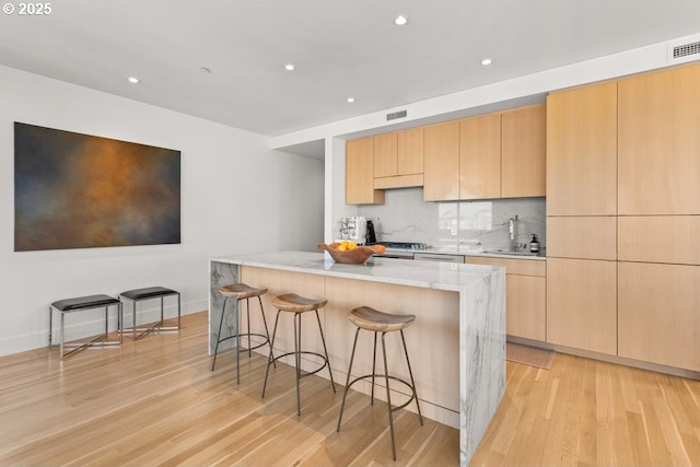 kitchen with a breakfast bar area, decorative backsplash, light stone countertops, light brown cabinets, and light hardwood / wood-style flooring