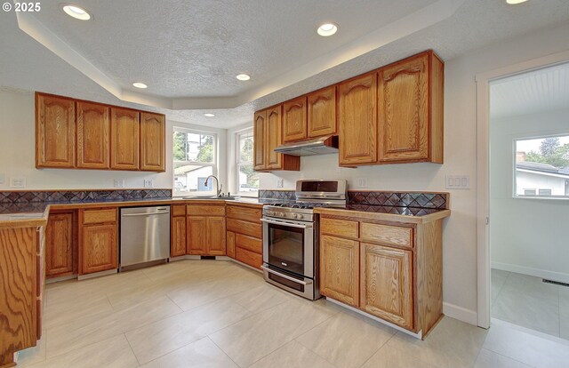 kitchen with appliances with stainless steel finishes, a sink, brown cabinets, a textured ceiling, and under cabinet range hood