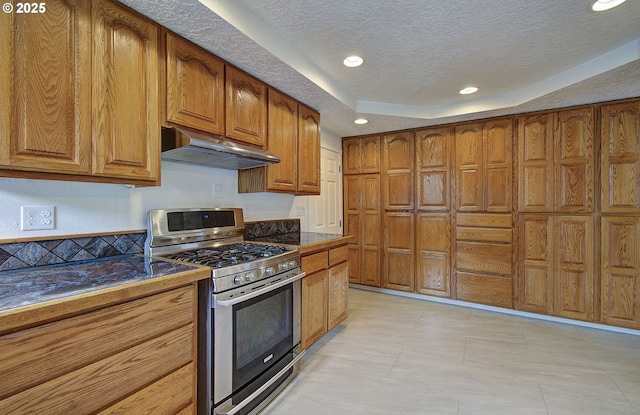 kitchen with stainless steel gas stove, under cabinet range hood, a tray ceiling, brown cabinets, and dark countertops