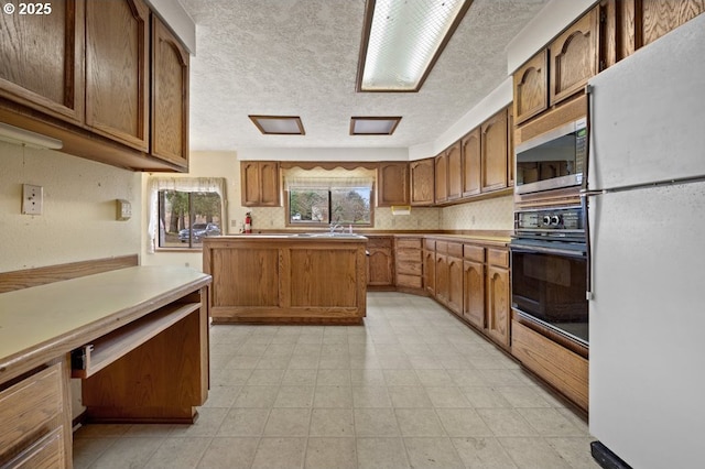 kitchen featuring a textured ceiling, stainless steel microwave, black oven, freestanding refrigerator, and brown cabinetry