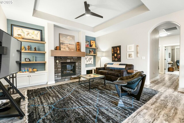 living room with arched walkways, a stone fireplace, a tray ceiling, and wood finished floors