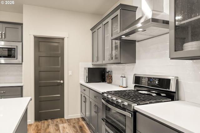 kitchen with gray cabinets, light countertops, light wood-style flooring, appliances with stainless steel finishes, and wall chimney exhaust hood