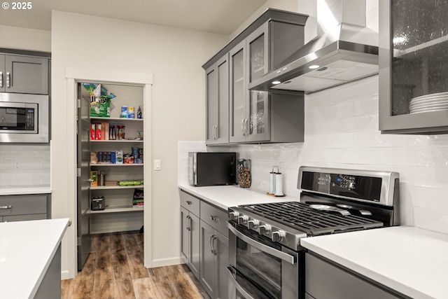 kitchen with wall chimney exhaust hood, appliances with stainless steel finishes, wood finished floors, light countertops, and gray cabinetry