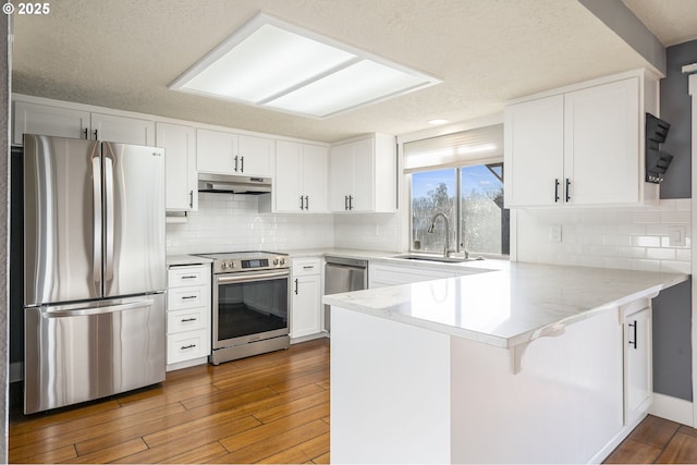 kitchen with a peninsula, a sink, hardwood / wood-style flooring, under cabinet range hood, and appliances with stainless steel finishes