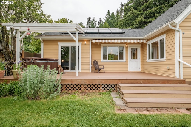 back of house with a hot tub, solar panels, a wooden deck, a shingled roof, and a lawn