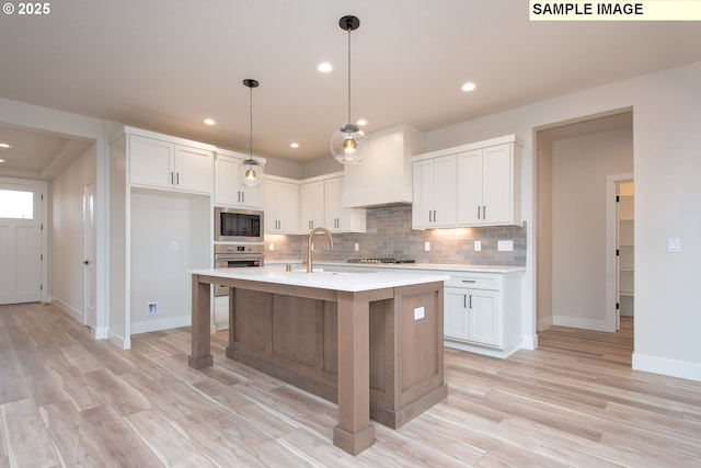 kitchen with appliances with stainless steel finishes, decorative light fixtures, premium range hood, white cabinets, and a center island with sink