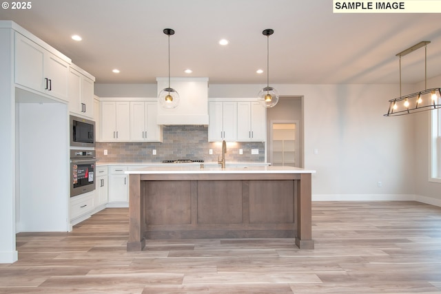 kitchen featuring hanging light fixtures, appliances with stainless steel finishes, premium range hood, white cabinets, and an island with sink