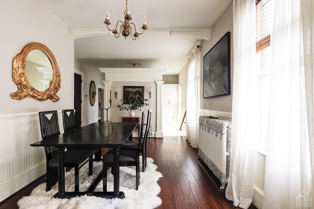 dining area featuring arched walkways, wainscoting, ornate columns, dark wood finished floors, and an inviting chandelier