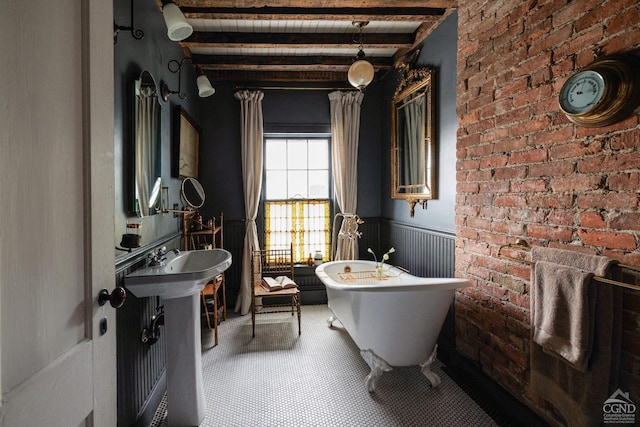 bathroom with beam ceiling, a freestanding bath, wainscoting, a sink, and brick wall