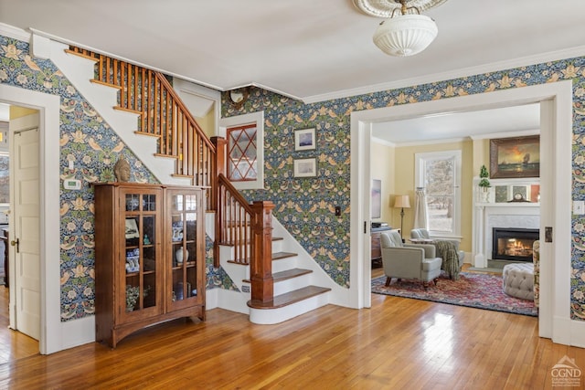 staircase featuring a glass covered fireplace, wood finished floors, crown molding, and wallpapered walls