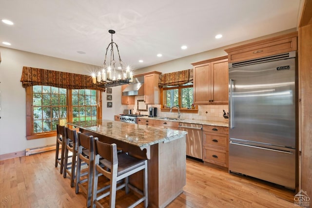 kitchen with pendant lighting, wall chimney range hood, light hardwood / wood-style flooring, appliances with stainless steel finishes, and a kitchen island