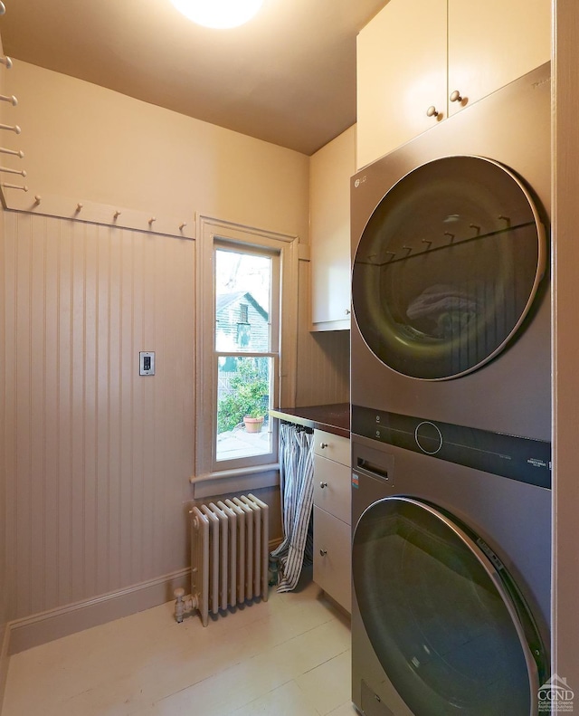 clothes washing area featuring cabinets, radiator heating unit, stacked washing maching and dryer, and wood walls