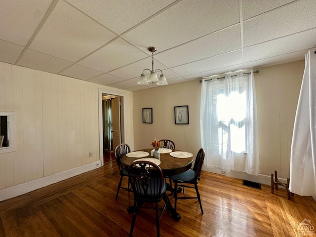 dining room with hardwood / wood-style floors, wooden walls, a drop ceiling, and a notable chandelier