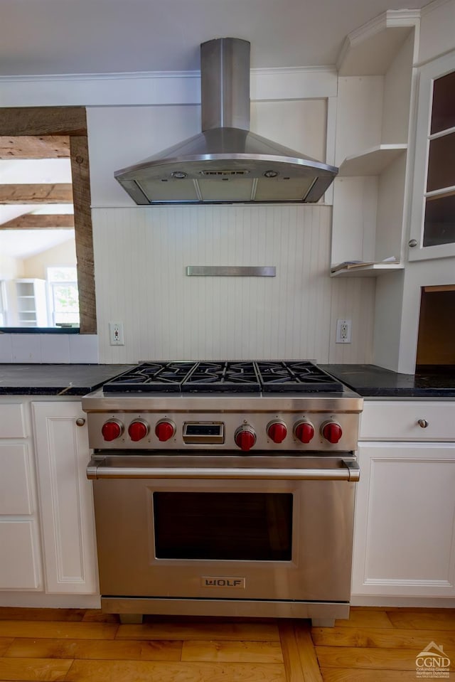 kitchen featuring wall chimney range hood, dark stone countertops, designer stove, light hardwood / wood-style floors, and white cabinetry