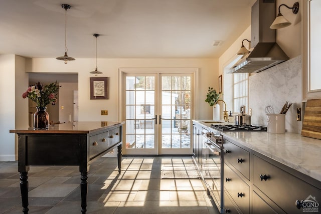 kitchen with wall chimney range hood, sink, high end stove, light stone countertops, and decorative light fixtures