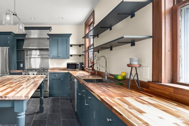 kitchen with wall chimney range hood, sink, stainless steel appliances, blue cabinets, and wood counters