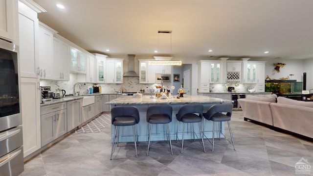 kitchen with stainless steel microwave, wall chimney range hood, light stone counters, an island with sink, and decorative light fixtures