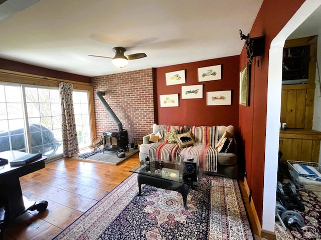 living room with wood-type flooring, a wood stove, ceiling fan, and brick wall