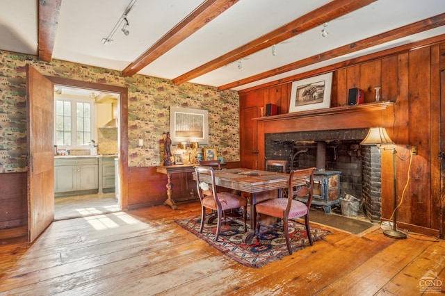 dining area featuring a wood stove, track lighting, wooden walls, light wood-type flooring, and beam ceiling