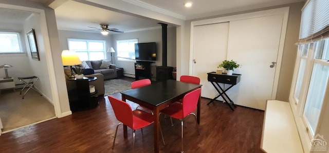 dining area featuring baseboards, a wood stove, a ceiling fan, crown molding, and dark wood-type flooring