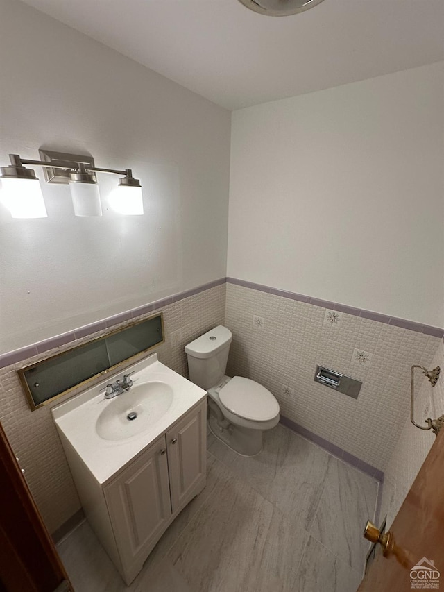 half bath featuring vanity, wainscoting, toilet, and tile walls