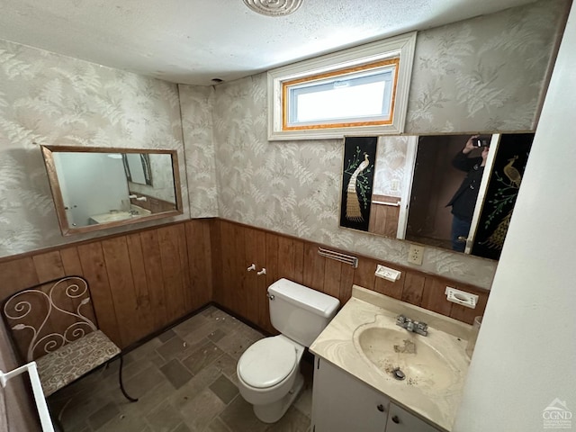 bathroom with wooden walls, vanity, toilet, a wainscoted wall, and wallpapered walls