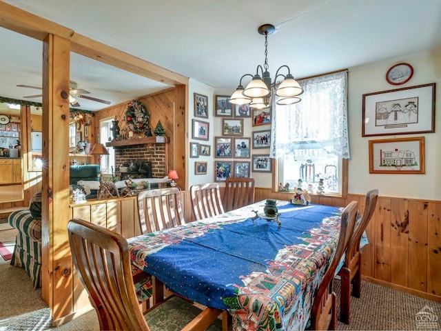 carpeted dining room with wooden walls, ceiling fan with notable chandelier, and a brick fireplace